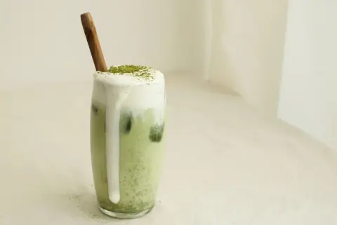 Un verre de matcha lait d'amande crémeux, garni de mousse et d'une pincée de poudre verte, avec une cuillère en bois.