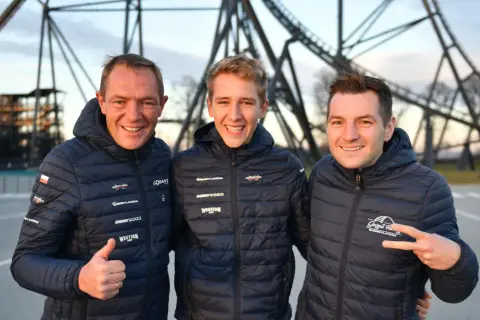 Trzech mężczyzn w kurtkach z logo Energylandii pozuje na tle konstrukcji roller coastera.