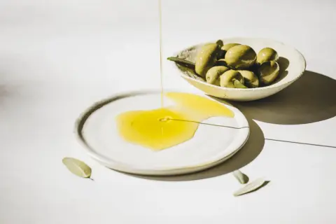 Flüssiges Olea europaea fruit oil fließt auf einen Teller, daneben eine Schale mit grünen Oliven und Blättern.