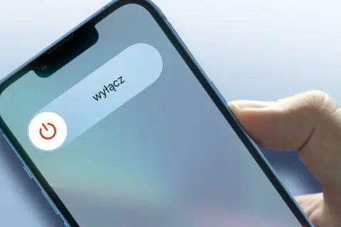 Dłoń trzyma iPhone'a z widocznym suwakiem "wyłącz" i ikoną zasilania.