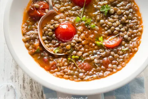 Una ciotola bianca piena di saporita zuppa di lenticchie con pomodorini e prezzemolo fresco. Un mestolo serve la zuppa.