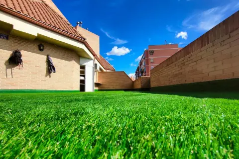 Un vibrante césped artificial para parques infantiles cubre una terraza con edificios de ladrillo y un cielo azul brillante.
