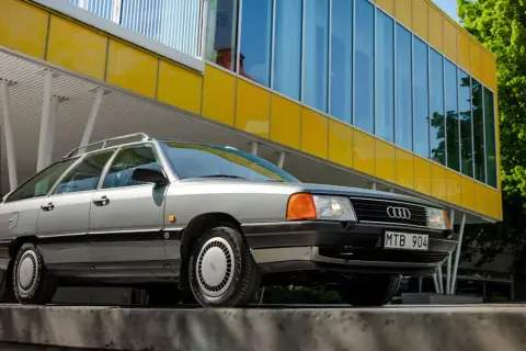 En grå Audi 100 C3 Avant står parkerad framför en modern gul byggnad med stora fönster.