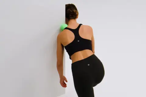 Femme utilisant une balle verte pour masser et muscler ses omoplates contre un mur blanc.
