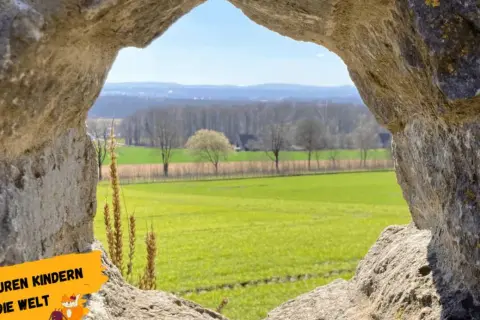 Ein Urlaub am Niederrhein durch ein steinernes Fenster: Grüne Felder, Bäume und ein blauer Himmel.