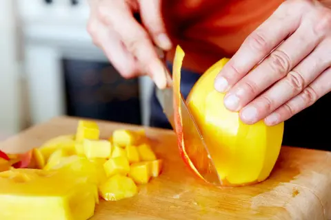 Hände schneiden eine reife Mango auf einem Holzbrett. Saftige Fruchtstücke liegen daneben.