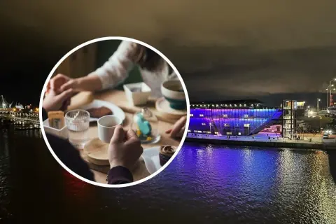 Para trzymająca się za ręce przy stoliku z kawą i ciastkiem, na tle nocnego Szczecina z oświetloną budowlą nad wodą.