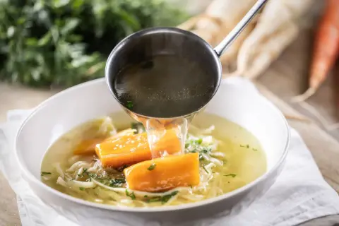 Un mestolo versa brodo caldo in una ciotola di zuppa di pollo con carote e tagliatelle.