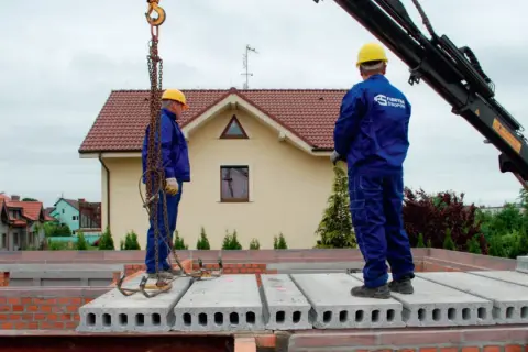 Dwóch budowlańców w żółtych kaskach montuje prefabrykowane płyty stropowe.