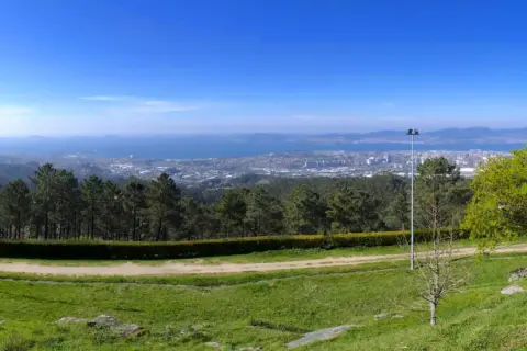 Vista panorámica desde el monte dos pozos, con un camino de tierra y césped en primer plano, árboles y un faro, y una ciudad y el mar al fondo.