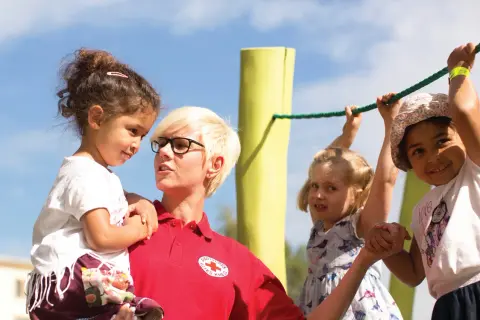 Frau vom Roten Kreuz fördert **Inklusion im Kindergarten**, indem sie ein Kind hält, während andere Kinder an einem Seil spielen.
