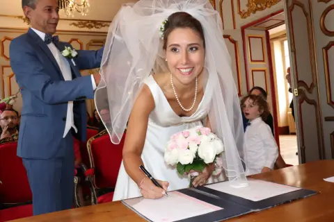 La mariée Anne-Claire Legendre signe son acte de mariage, bouquet à la main. Un enfant aux boucles blondes observe la scène.