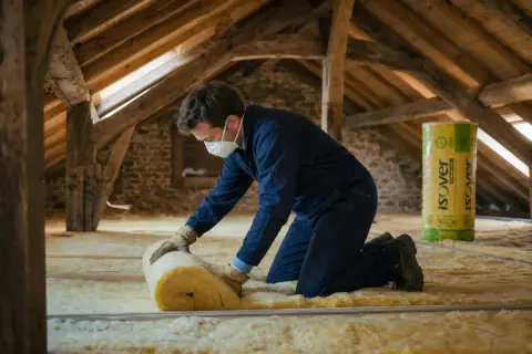 Un ouvrier installe un isolant jaune dans un grenier. Le rouleau d'isolant et la mention "prix isolation toiture" sur le produit suggèrent une rénovation pour améliorer le confort thermique.