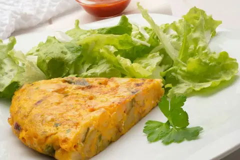 Una porción de tortilla de patatas, dorada y jugosa, acompañada de una fresca ensalada verde y un toque de perejil.