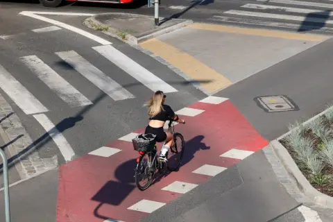 Kobieta na rowerze przejeżdża przez czerwony pas rowerowy, zbliżając się do **uwaga przejście dla pieszych**.