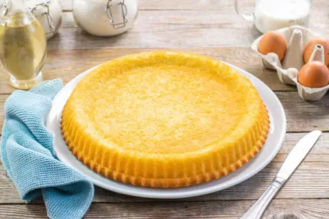 Una torta furba dorata, appena sfornata, su un piatto bianco. Ingredienti come uova e olio sono visibili sullo sfondo.