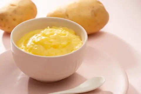 Ein Schälchen mit cremigem Kartoffelbrei, bereit für ein Baby. Zwei Kartoffeln liegen im Hintergrund.
