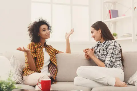 Dwie kobiety siedzą na kanapie, rozmawiając. Jedna z nich gestykuluje, pokazując różne techniki komunikacji interpersonalnej.