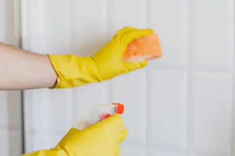Ręce w żółtych rękawiczkach czyszczą białe płytki. Jedna ręka trzyma gąbkę, druga spryskiwacz. Idealne rozwiązanie, czym wyczyścić białe fugi.