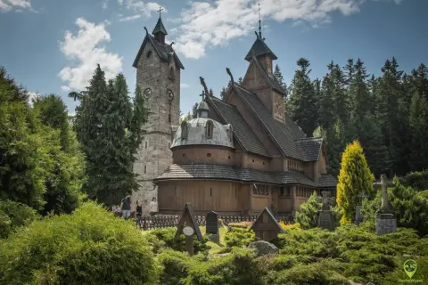 Drewniany kościółek i kamienna wieża z zegarem, otoczone zielenią i nagrobkami. Jedna z głównych karpacz atrakcji.