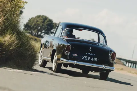 Svart Simca Aronde glider längs en kustväg, en klassisk skönhet bland gräs och träd.