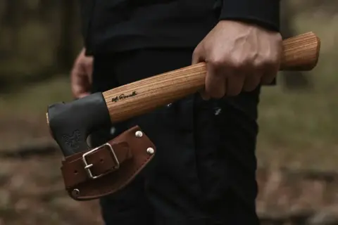 Mano sujetando hachas tácticas con funda de cuero. El mango de madera tiene una inscripción.