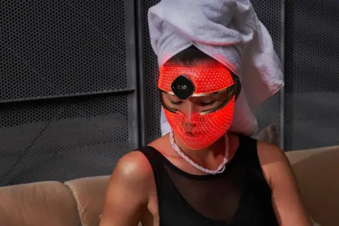 Una mujer con toalla en la cabeza usa una máscara LED facial roja. Descubre para qué sirve esta máscara de belleza.