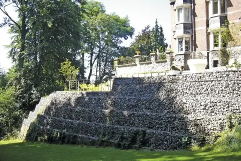 Imponująca skarpa na granicy działki, zbudowana z kamieni w gabionach, tworzy tarasy porośnięte zielenią. Na górze widać fragment rezydencji.