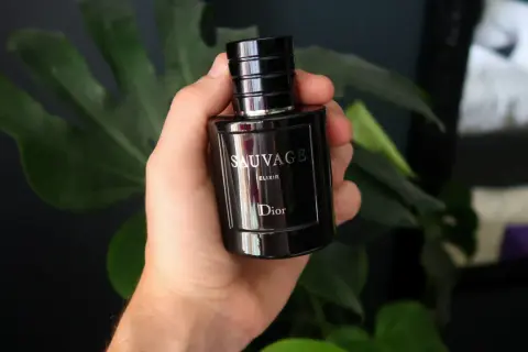 Dłoń trzyma flakon Dior Sauvage Elixir 60ml, otoczony zielonymi liśćmi.