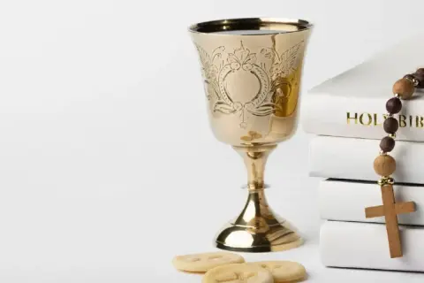 Złoty kielich, hostie, różaniec i stos białych ksiąg, w tym "Holy Bible". Symbolika sakramentu, jak akt chrztu.