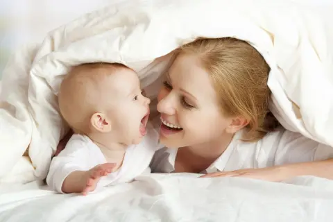Mutter und ihr **baby 5 wochen** alt kuscheln unter einer Decke und lachen sich an.