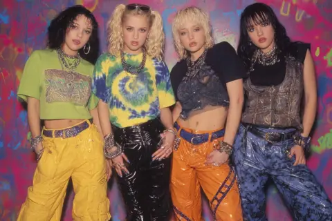Vier junge Frauen im coolen **mode 90er jahre** Stil mit bunten Outfits und auffälligem Schmuck posieren vor einer Graffiti-Wand.