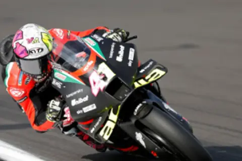 Piloto de MotoGP inclinado en una curva, con su casco blanco y rosa y la moto negra con detalles amarillos.
