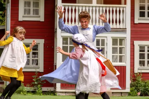 Barn leker vid Junibacken Madicken hus. En vuxen i blå klänning och två barn i tidsenliga kläder.