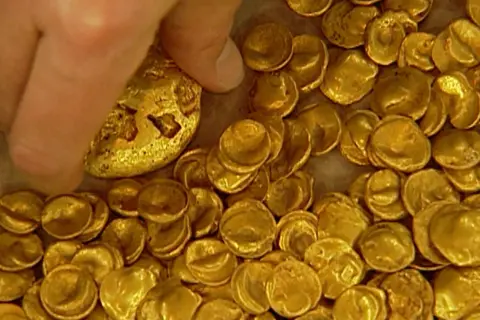 Ein Finger berührt einen Teil des Goldschatzes von Manching, der aus vielen kleinen, unregelmäßigen Goldmünzen besteht.