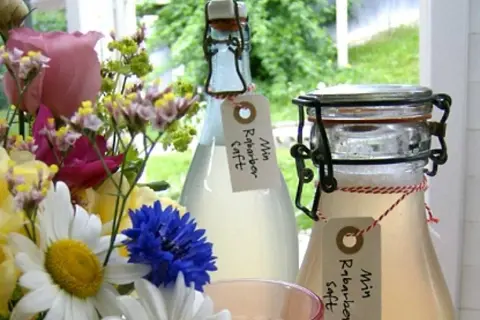 Två flaskor hemgjord rabarbersaft står bredvid en blomsterbukett. Etiketter på flaskorna lyder "Min Rabarbersaft".