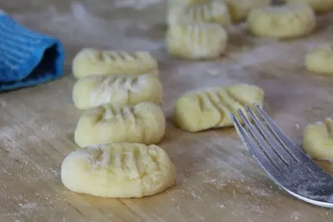 Frisch geformte Gnocchi liegen auf bemehltem Holz. Eine Gabel und ein blauer Lappen deuten darauf hin, dass das Gnocchi selber machen gerade erst begonnen hat.