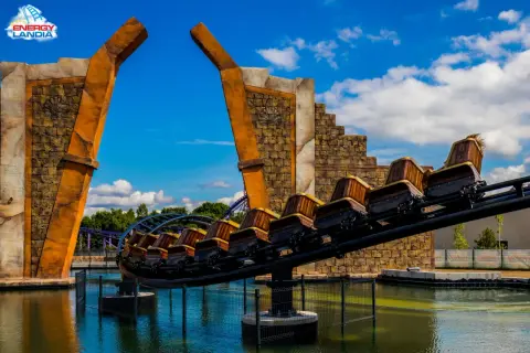 Ekipa rollercoastera w Energylandii gotowa na przygodę! Drewniane wagoniki pędzą nad wodą, a za nimi widać fioletowe tory.