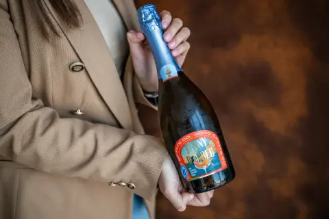 Kobieta w beżowym żakiecie trzyma butelkę musującego napoju, idealnego na międzynarodowy dzień prosecco.