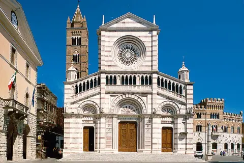 Il Duomo di Grosseto, con la sua facciata a strisce bianche e rosa, domina la piazza. Accanto, un campanile medievale si staglia contro il cielo azzurro.