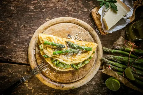Pyszny omlet ze szparagami, posypany parmezanem, czeka na widelcu. Obok świeże szparagi i kawałek sera.