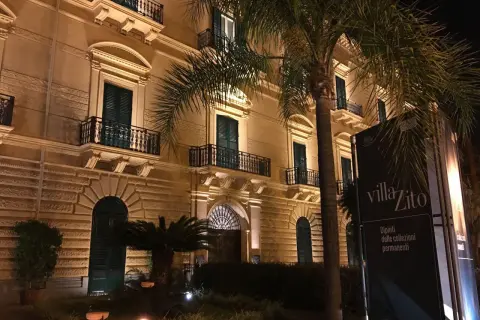Villa Zito a Palermo, un'elegante dimora illuminata di notte, con palme che incorniciano la facciata e un cartello che annuncia "Dipinti delle collezioni permanenti".