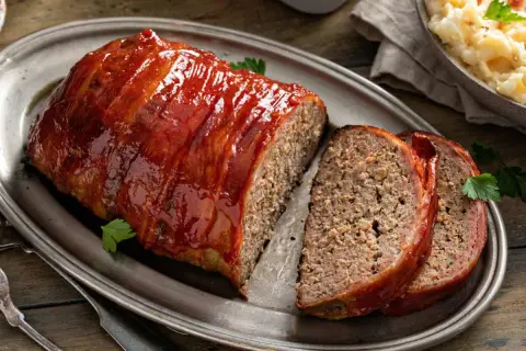 Un jugoso pastel de carne cubierto de glaseado rojo brillante, envuelto en tocino y servido en una bandeja de plata.