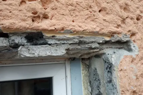 Uszkodzony montaż nadproża w starym domu, widoczne zbrojenie i luźne elementy betonowe nad oknem.