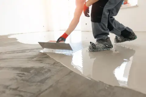 Obrero con guantes rojos y calzado especial nivela un suelo con mucho desnivel usando una llana dentada.