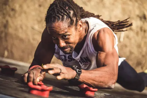 Man with dreadlocks on a climbing wall, his forearm punch showing determination as he grips the holds.