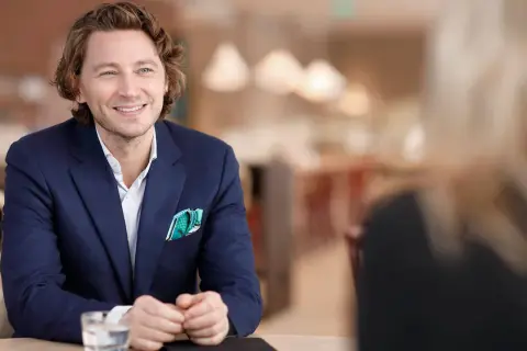 Rasmus Nerman, klädd i en mörkblå kavaj, ler och tittar åt sidan under ett samtal vid ett bord.