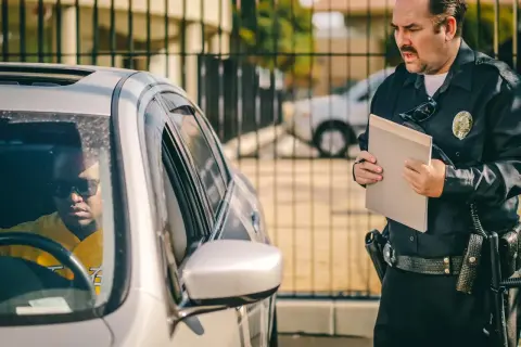 Policjant konfrontuje kierowcę po ucieczce z miejsca kolizji. Mężczyzna w okularach przeciwsłonecznych siedzi w samochodzie, podczas gdy funkcjonariusz trzyma teczkę.