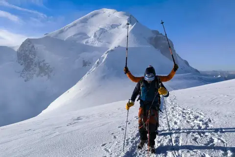 Un alpiniste victorieux lève ses bâtons sur fond de Mont Blanc. L'ascension est réussie, la neige brille sous le ciel bleu.