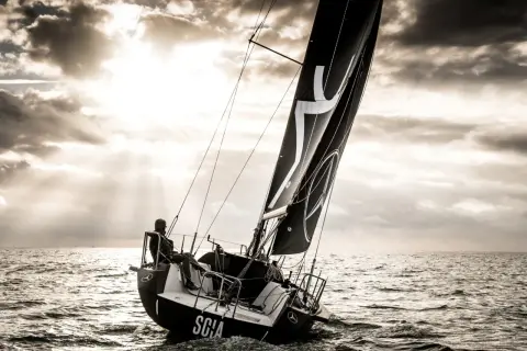 Vela nera con logo, un uomo a bordo di una barca a vela che naviga in mare con un cielo drammatico. L'imbarcazione mostra un angolo di deriva accentuato.